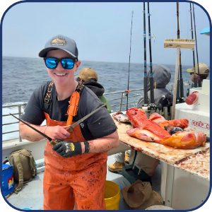 Person in orange overalls on boat, smiling with fish and fishing rods nearby.