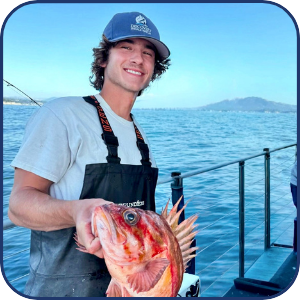 Person on a boat holding a fish with ocean and distant land in background.