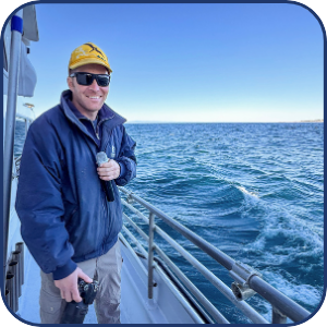 Man in yellow cap on boat holding a camera, with ocean in background.