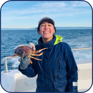 Person smiling on a boat holding a crab, with ocean in the background.