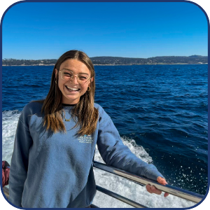 Smiling person with glasses on a boat, blue ocean background, clear sky.