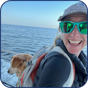 Person in sunglasses smiling on a boat with a dog, ocean in the background.