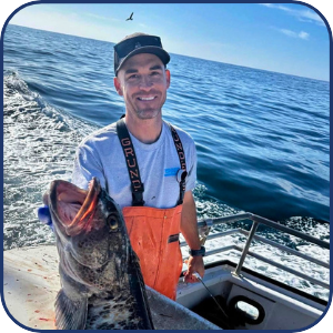 Man on boat holding a large fish, smiling, with ocean in background.